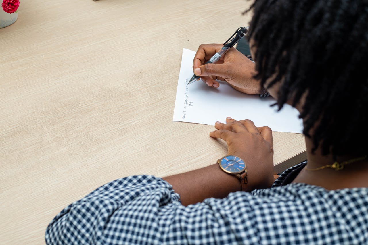 An individual writing notes on paper at a wooden desk in Lagos, Nigeria, wearing a wristwatch.